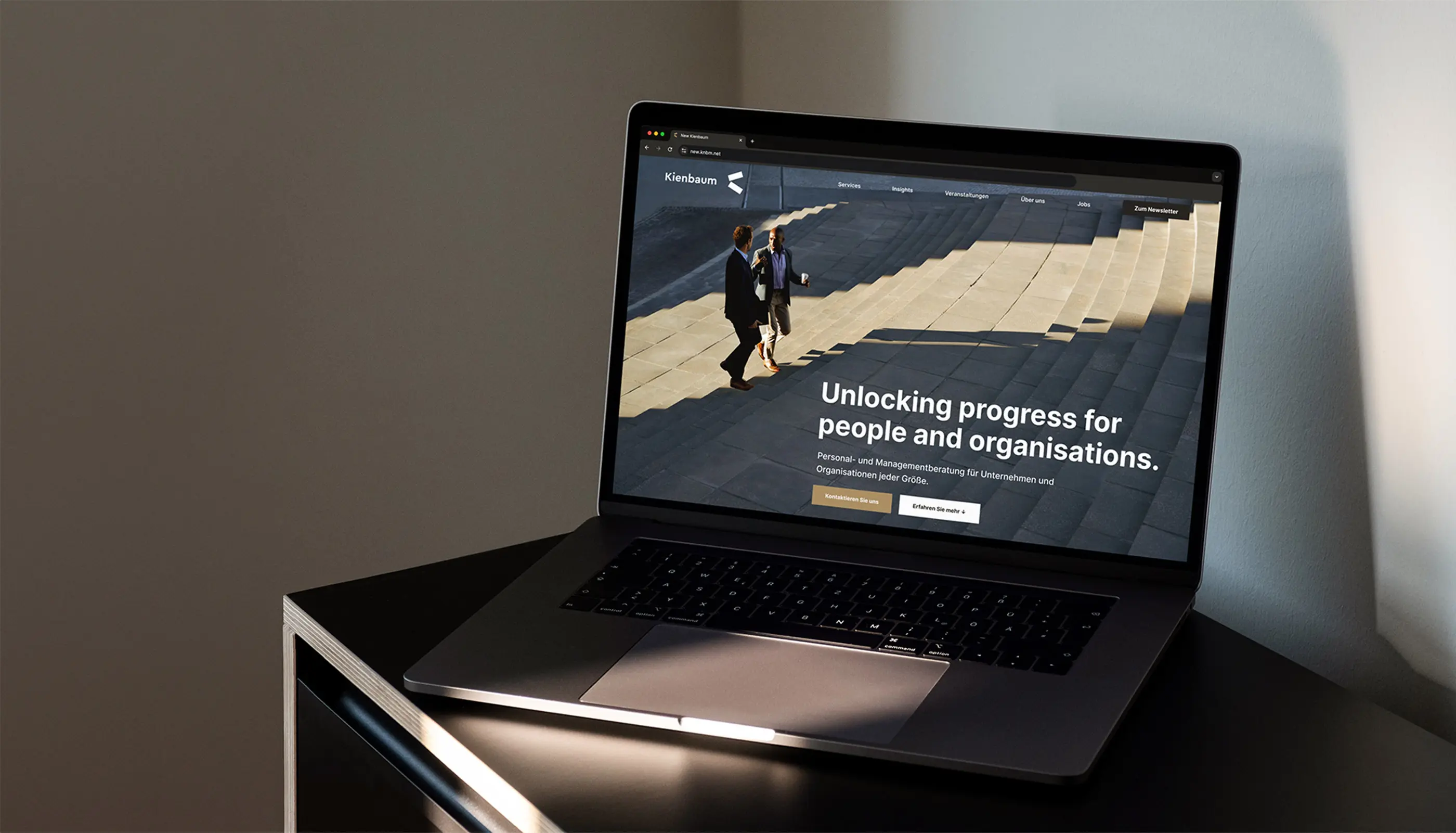 Laptop on a black table displaying a website homepage with two men talking on stairs and the text 'Unlocking progress for people and organisations.'