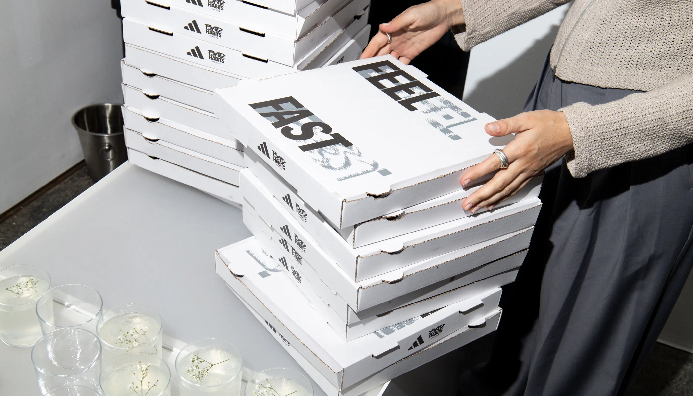 Person stacking white pizza boxes with 'FEEL FAST' printed on the top box on a table next to glasses of clear drinks with decorative sprigs.
