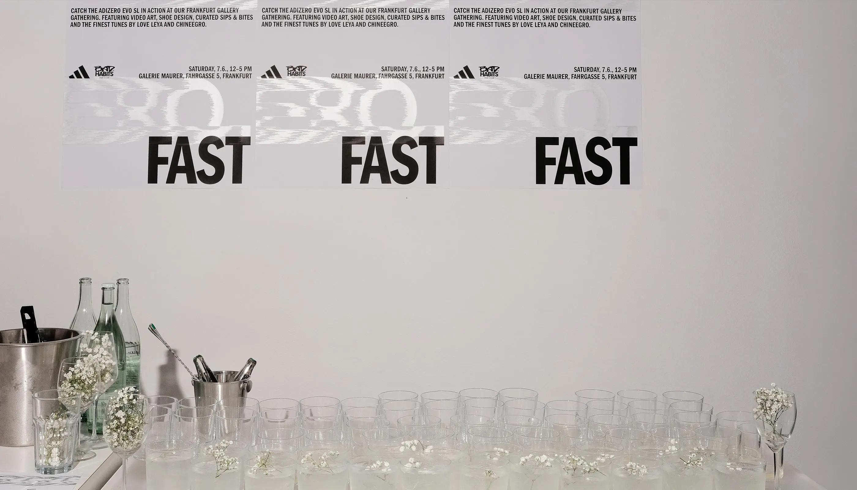 Three posters on a wall with 'ADIZERO FAST' text promoting an event at Galerie Maurer, Frankfurt; below a table with glasses of water garnished with small white flowers, bottles, and ice buckets.