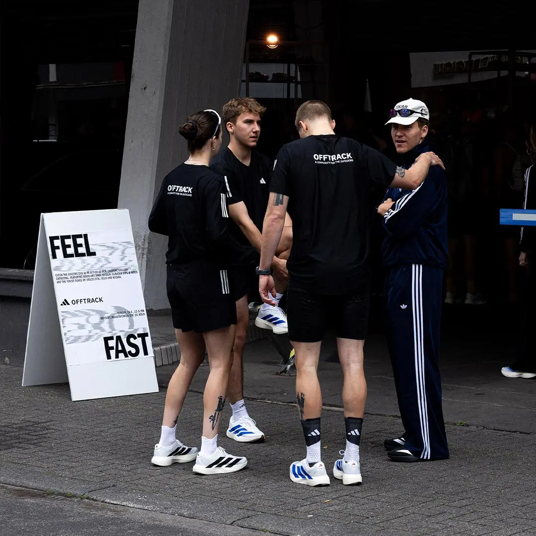 Group of four people standing outside, three wearing black OFFTRACK shirts and Adidas shoes, and one in a navy tracksuit and white cap.