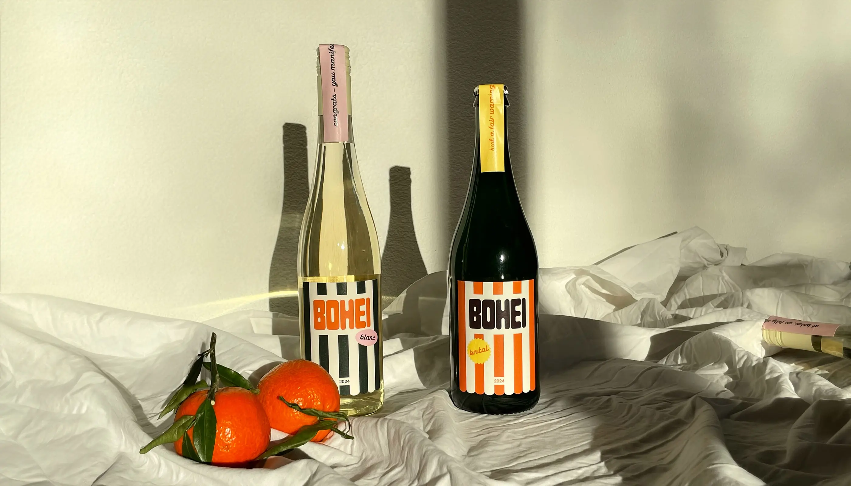 Two bottles of Bohei wine, one blanc with black and white stripes and one brutal with orange and white stripes, placed on white fabric next to three oranges with green leaves.