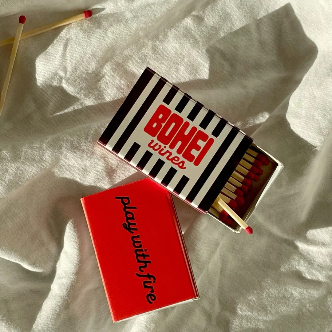 Two matchboxes on a white fabric; one open with red-tipped matches visible and striped black and white design reading 'BOHEI wines', the other closed and red with the text 'play with fire'.