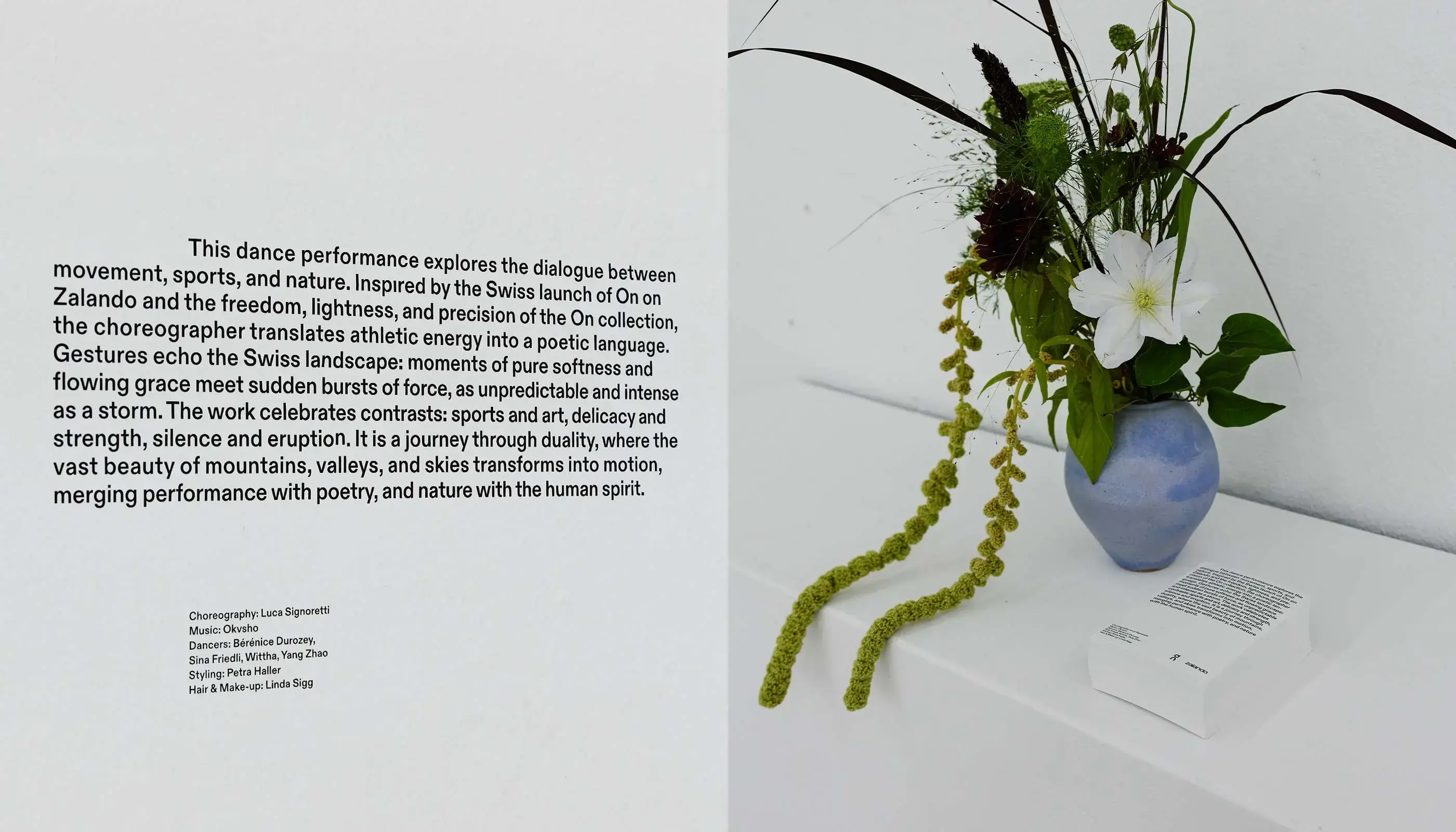 Blue ceramic vase with a floral arrangement including a white flower and green foliage, next to a folded paper on a white surface against a white wall.