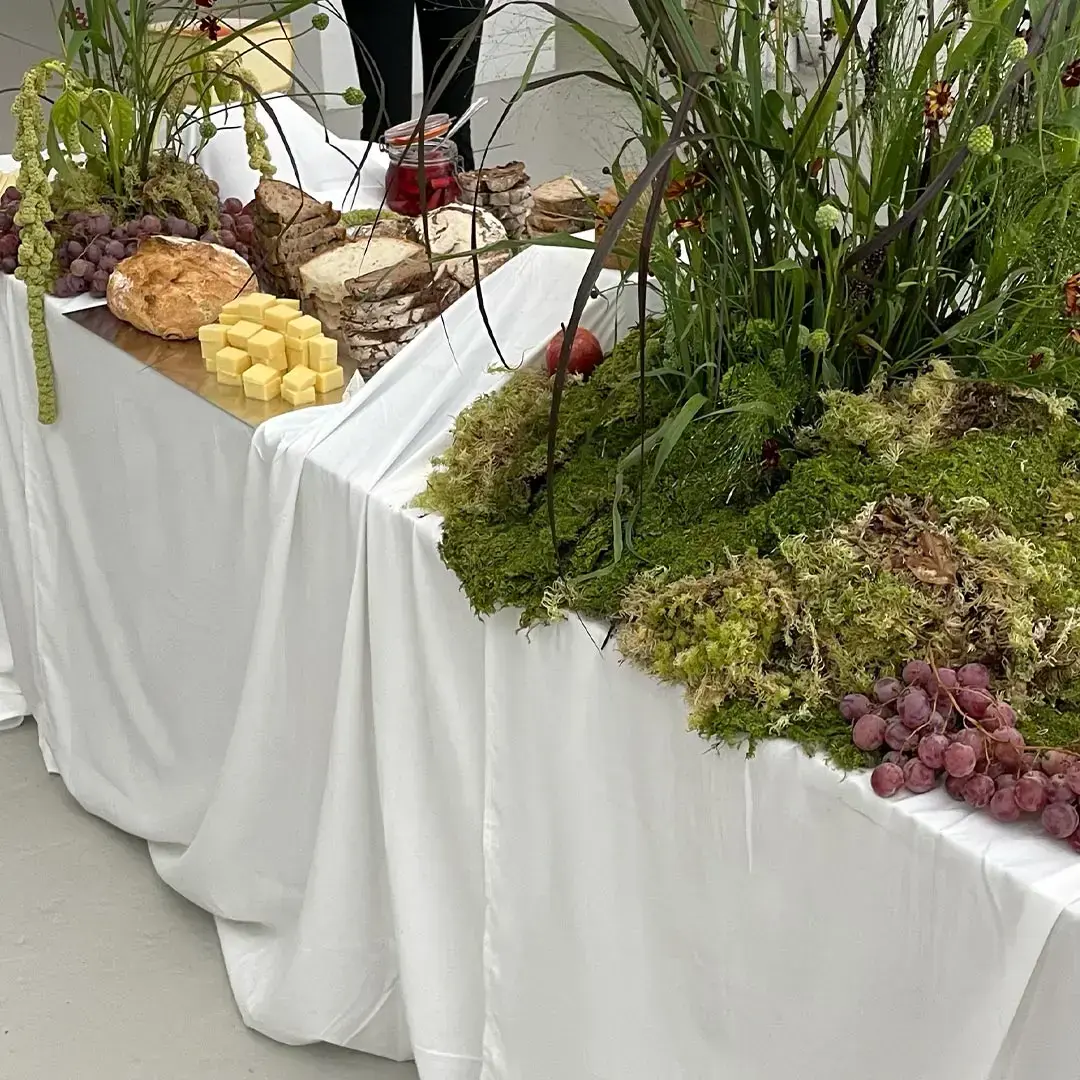 Table draped in white cloth displaying sliced bread, cubed cheese, grapes, a round loaf, glass jar of red jam, and green moss with tall wild plants.