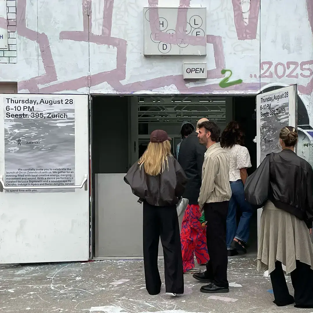 People entering a gallery or event space with open doors displaying posters about a Thursday, August 28 event in Zurich.